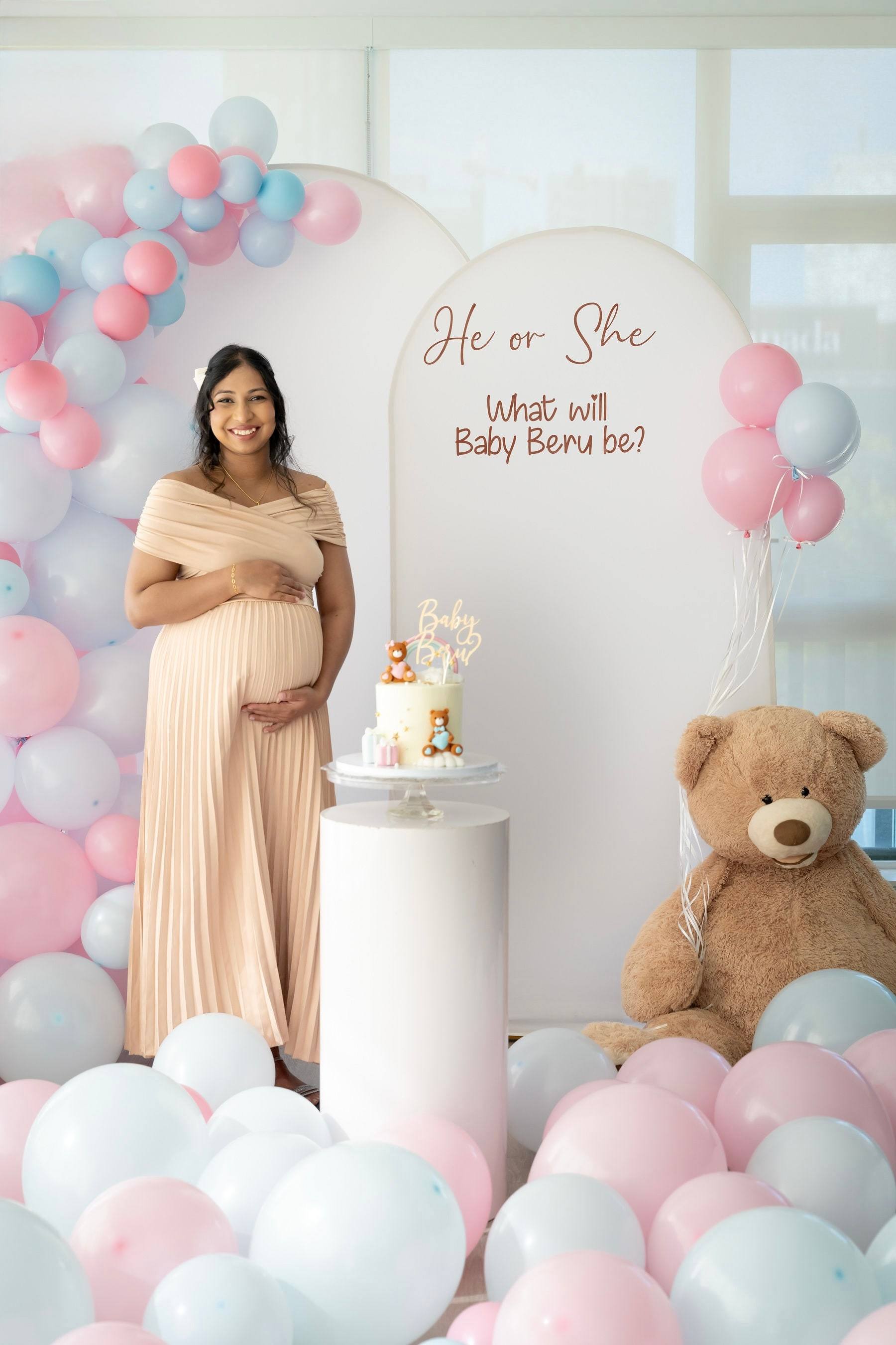 Mother-to-be beside the cake and teddy bear under a balloon arch