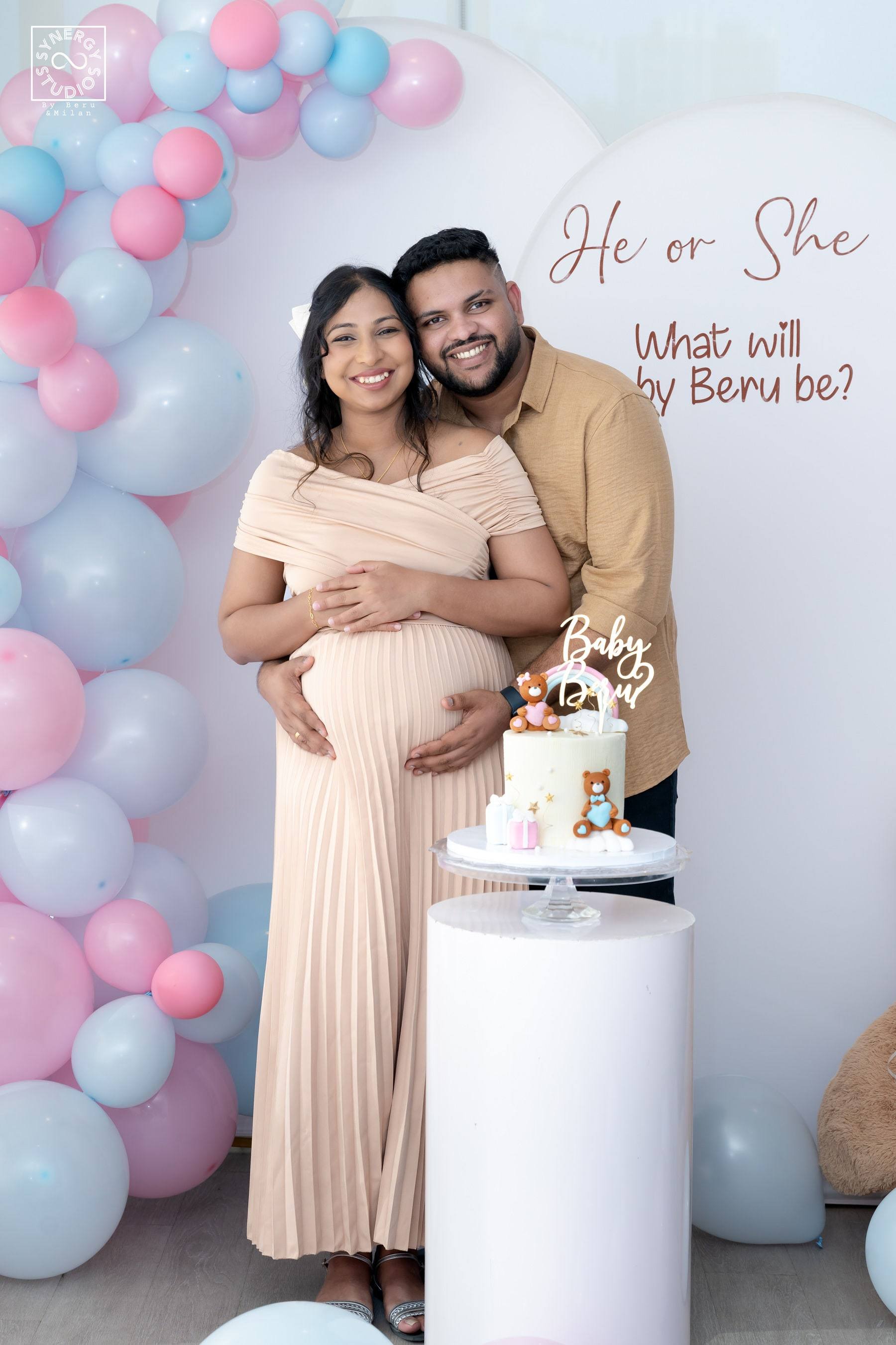 Parents-to-be embracing in front of a pastel balloon arch and "he or she" sign