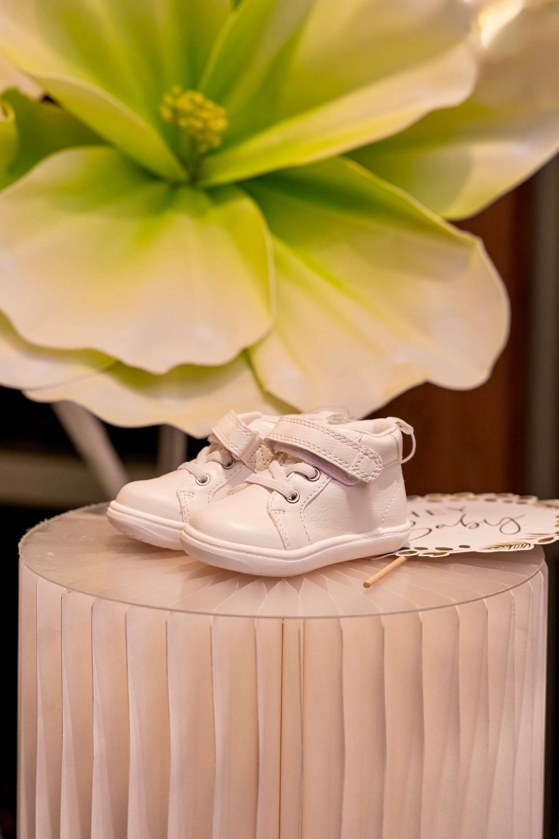 Tiny baby shoes displayed on a pink pedestal beside a paper flower