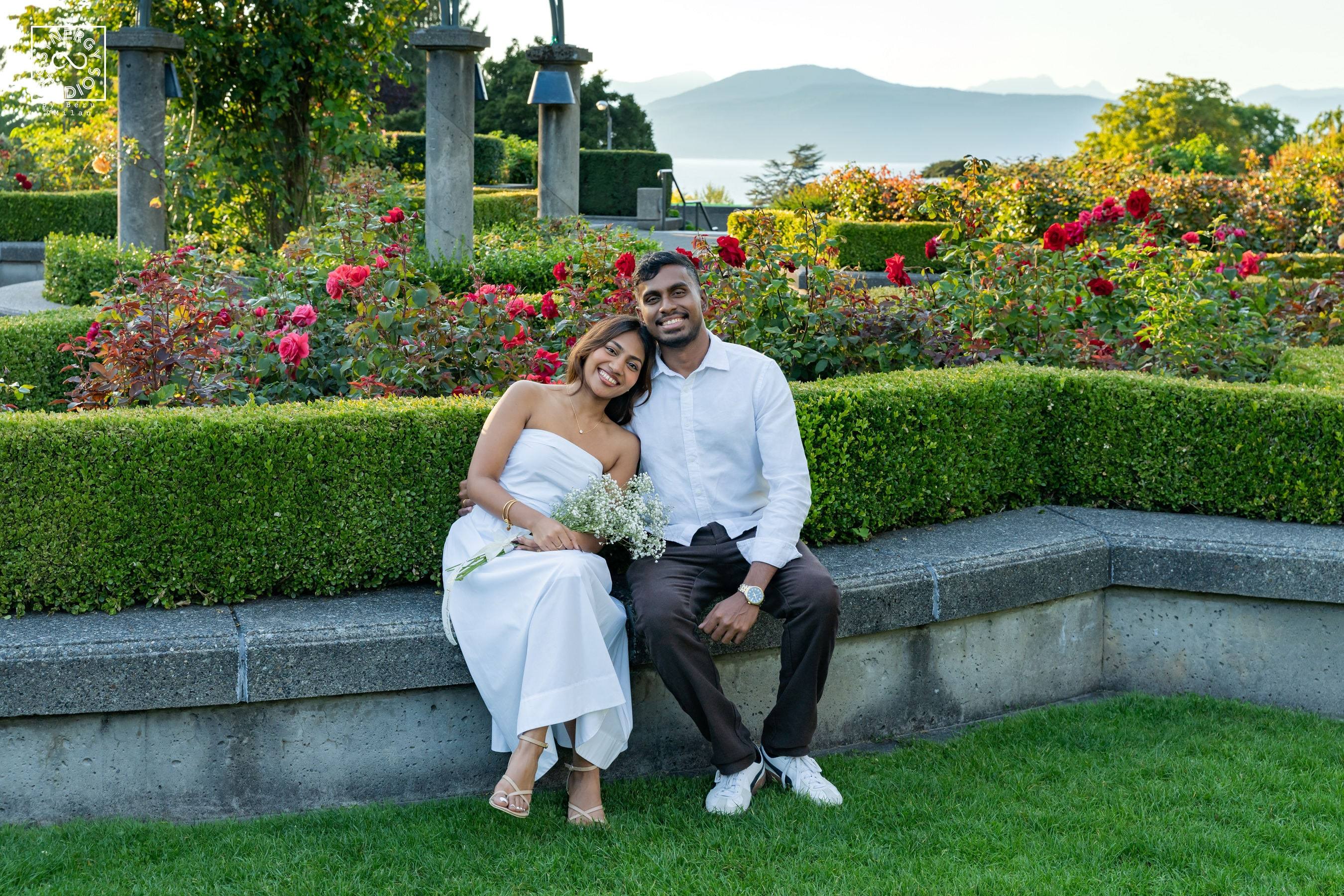 Couple sitting together on a stone ledge in a rose garden with mountains and the ocean behind them