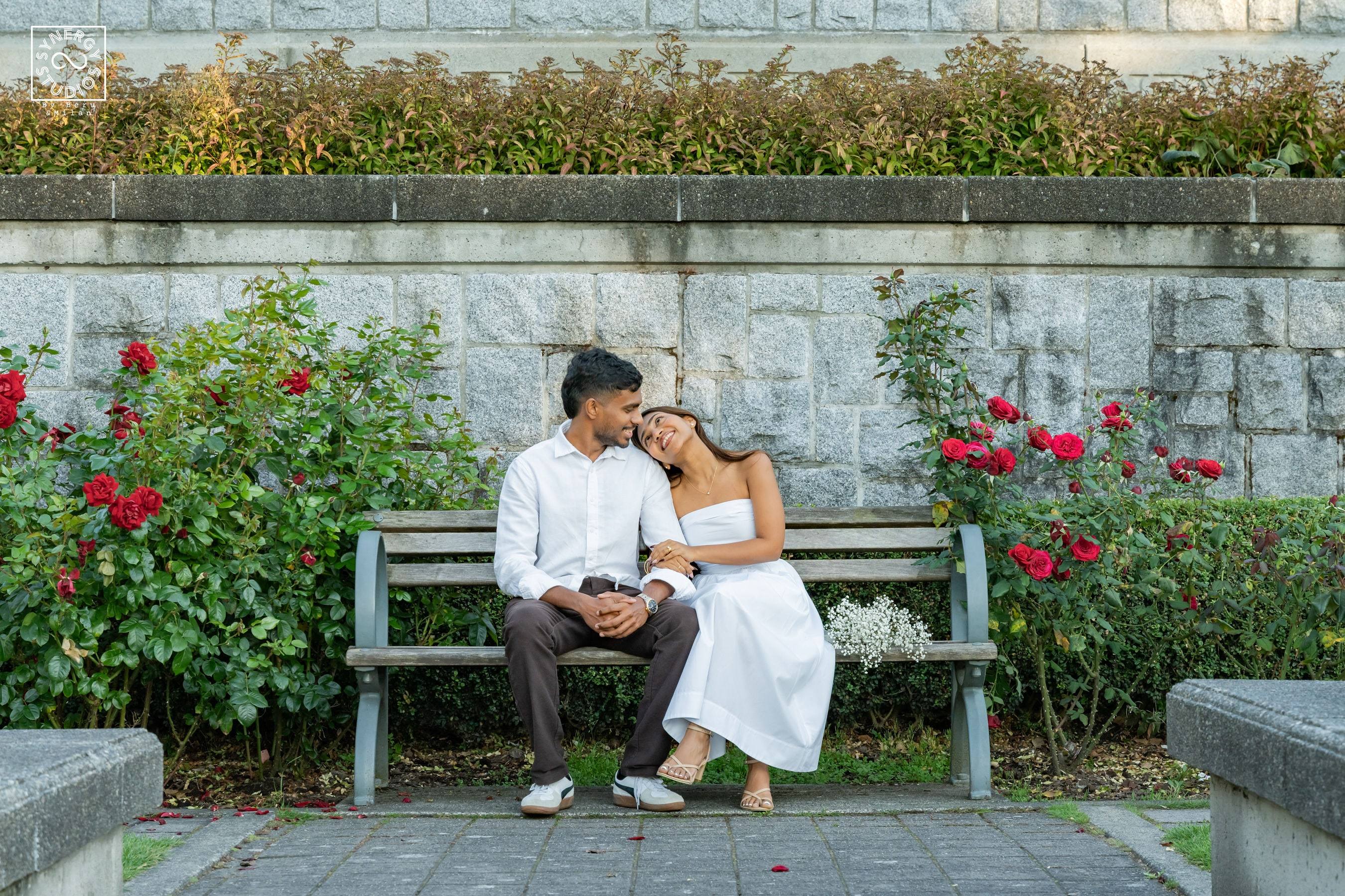 Couple leaning into each other on a wooden bench framed by red roses and a stone wall