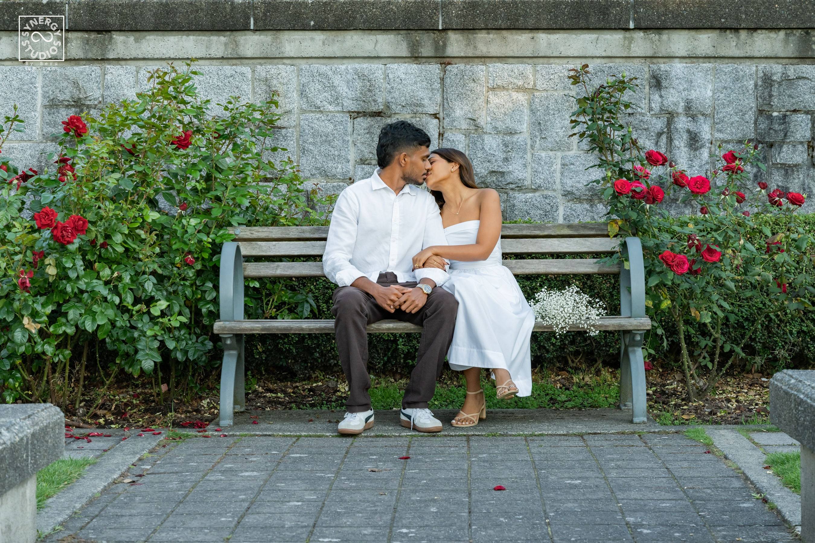 Couple sharing a kiss on a garden bench surrounded by red rose bushes