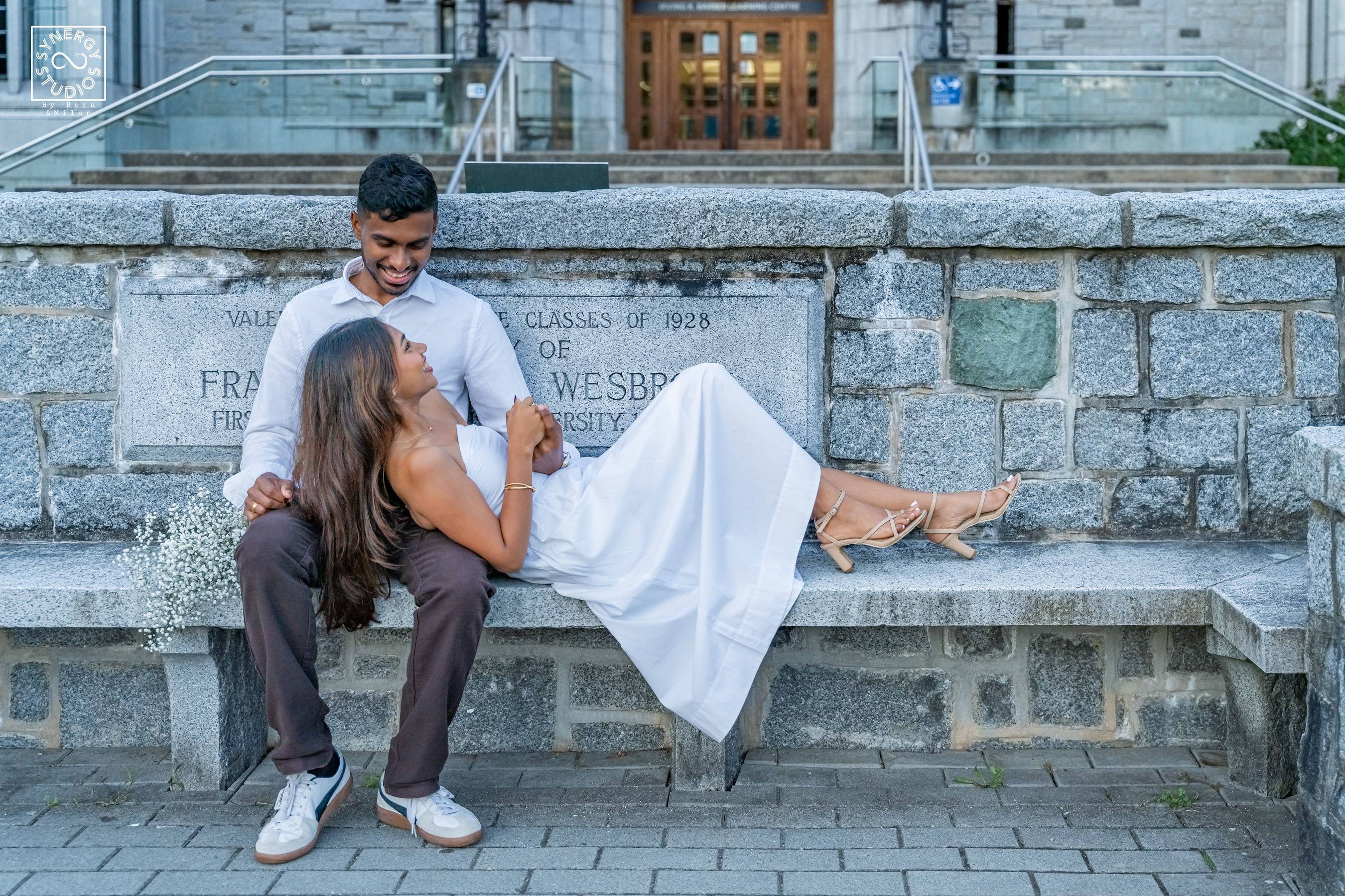Woman in a white dress lying across her partner's lap on a stone bench outside a heritage building