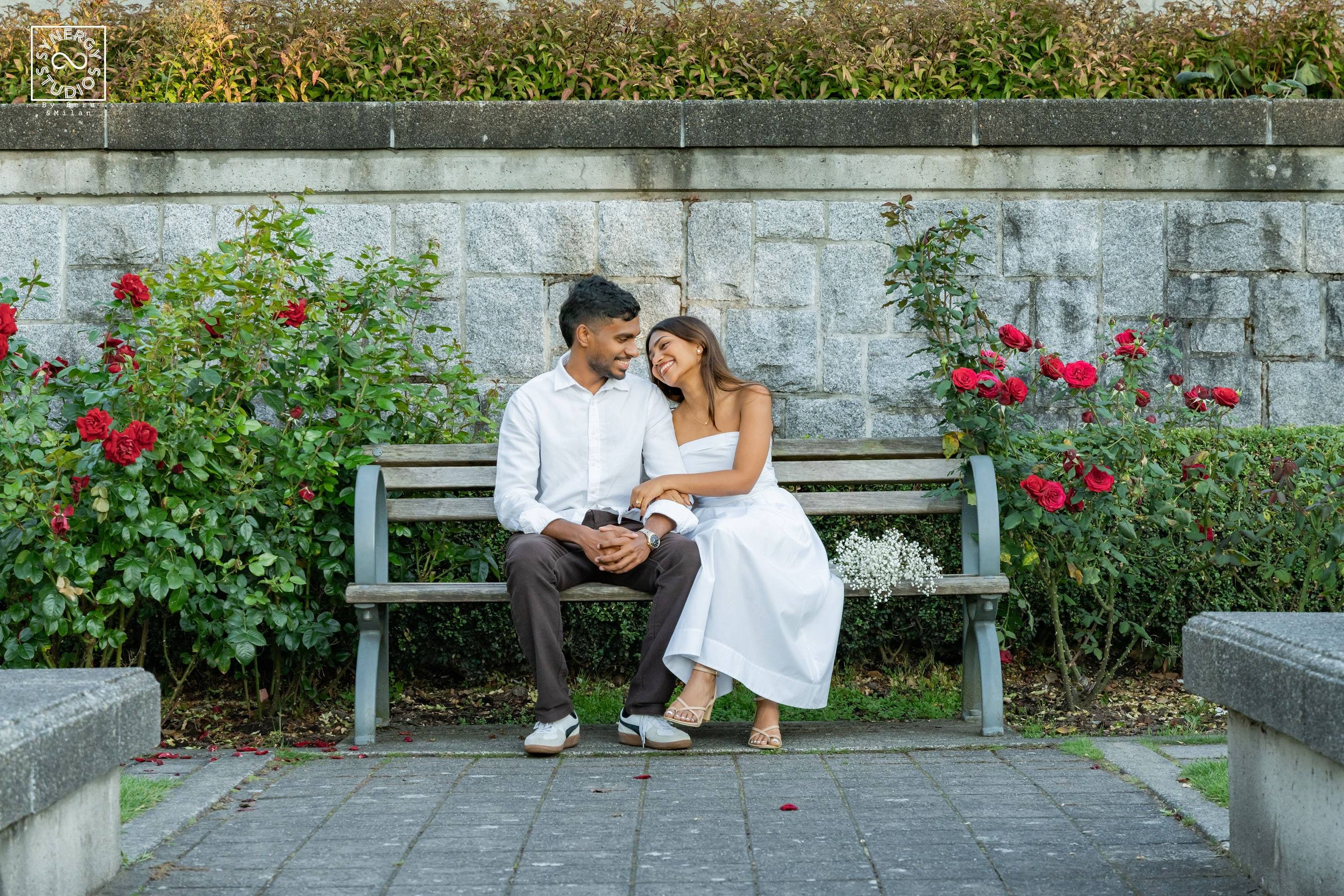 Couple seated on a bench between red roses with a stone wall behind
