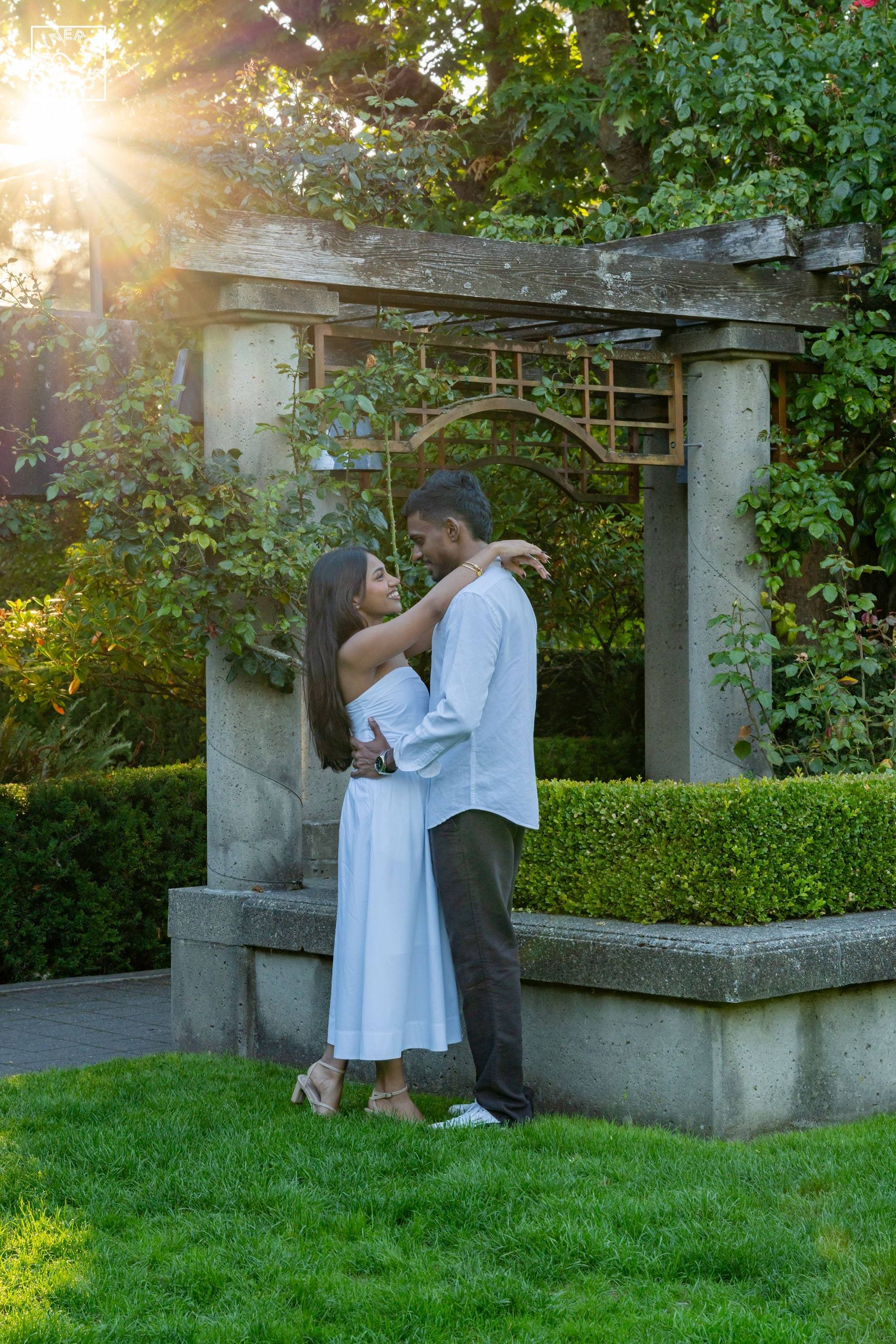 Couple embracing under a stone pergola as the sun flares through the trees
