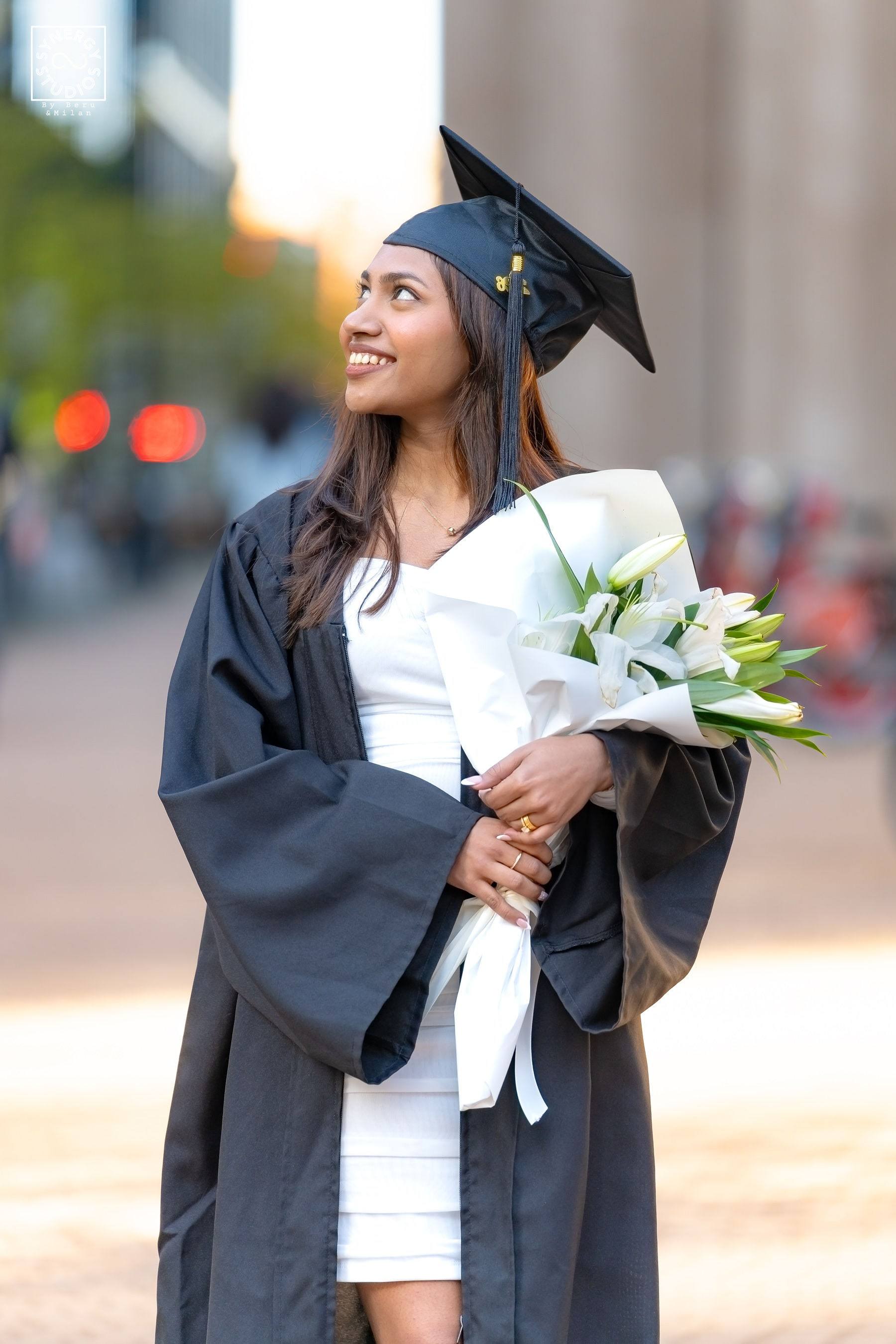 Graduate in a black gown holding a bouquet of white lilies on campus
