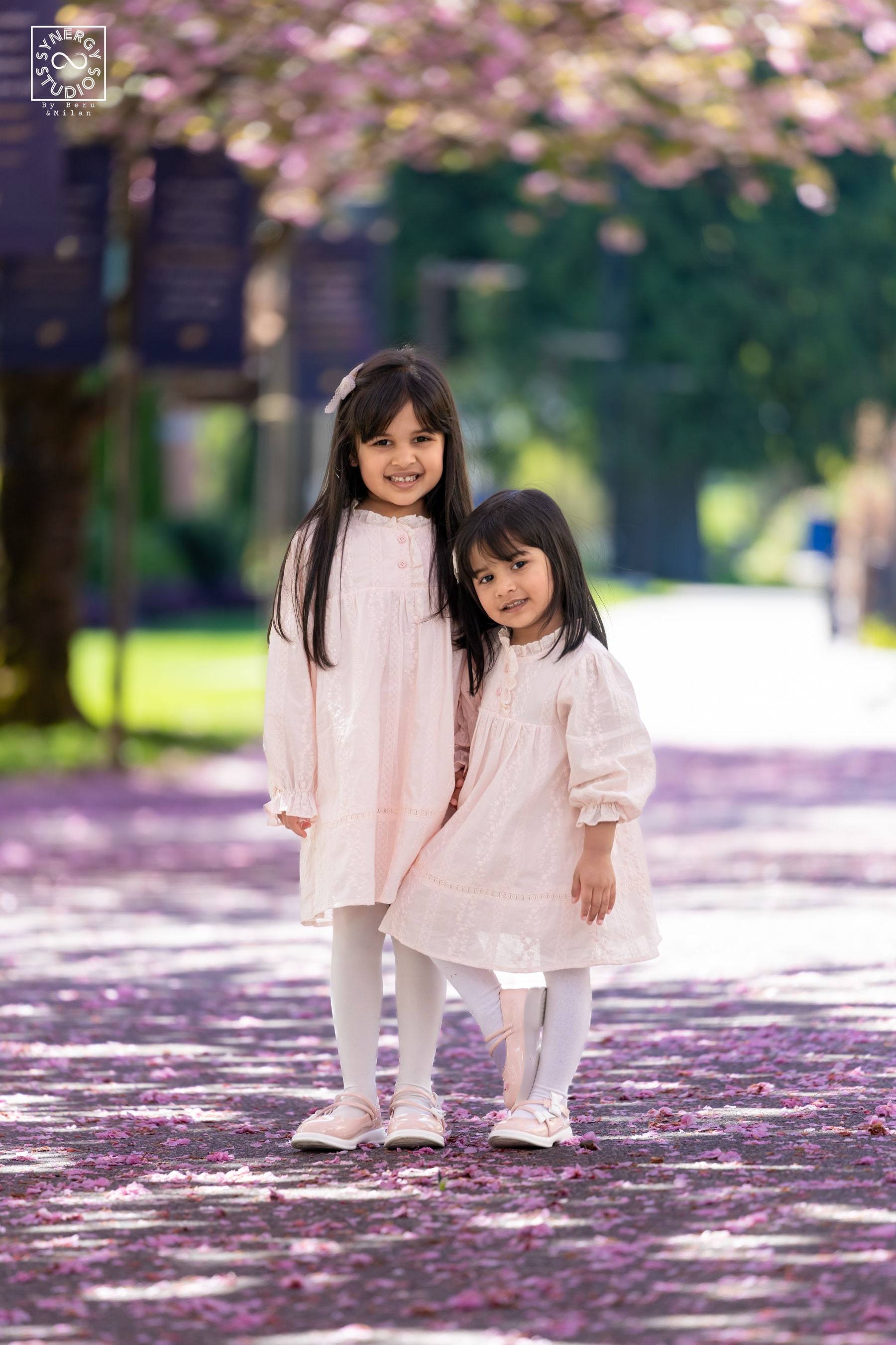Two young sisters in cream dresses standing under cherry blossom trees