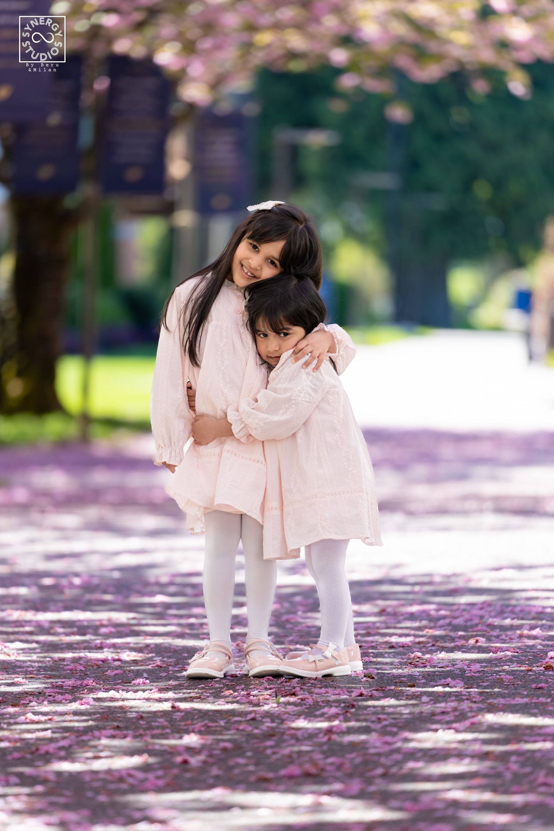 Little sisters hugging on a path covered in pink cherry blossom petals