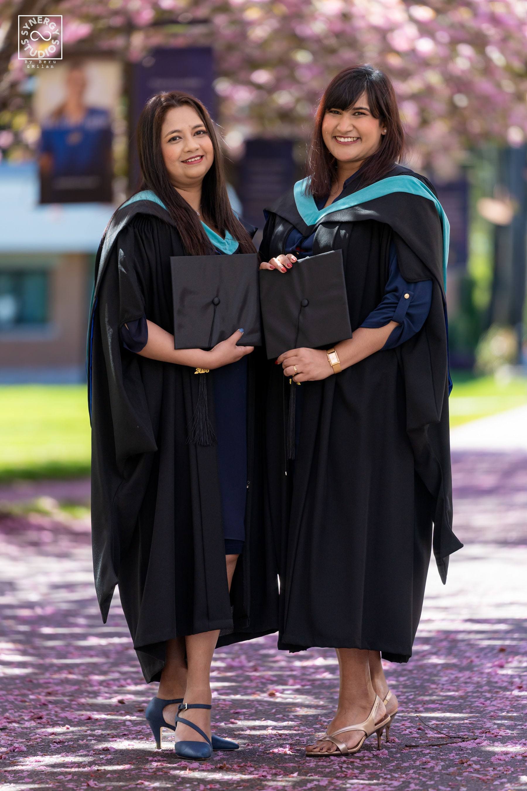 Two graduates in master's hoods smiling together under the blossoms