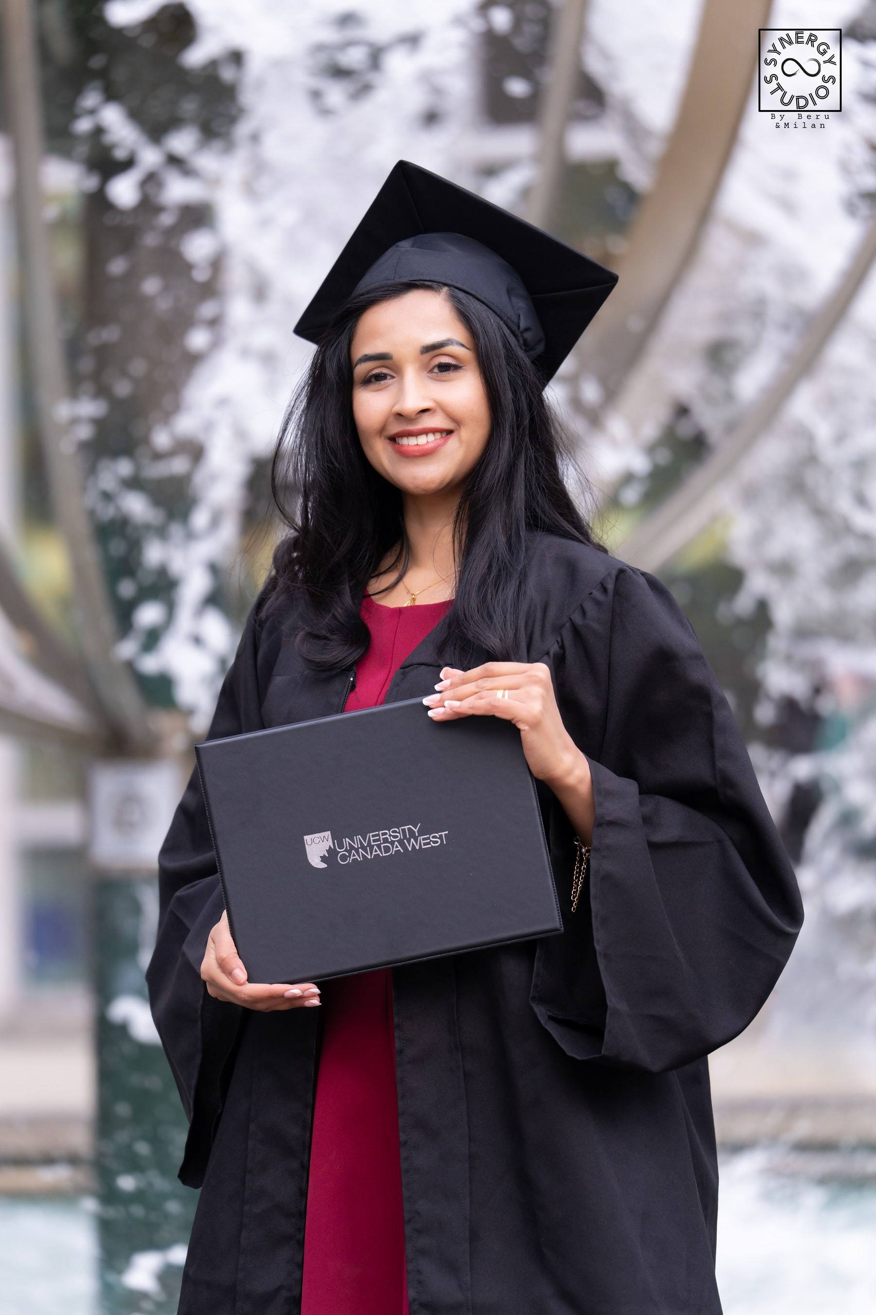 Graduate in cap and gown holding her diploma in front of a fountain