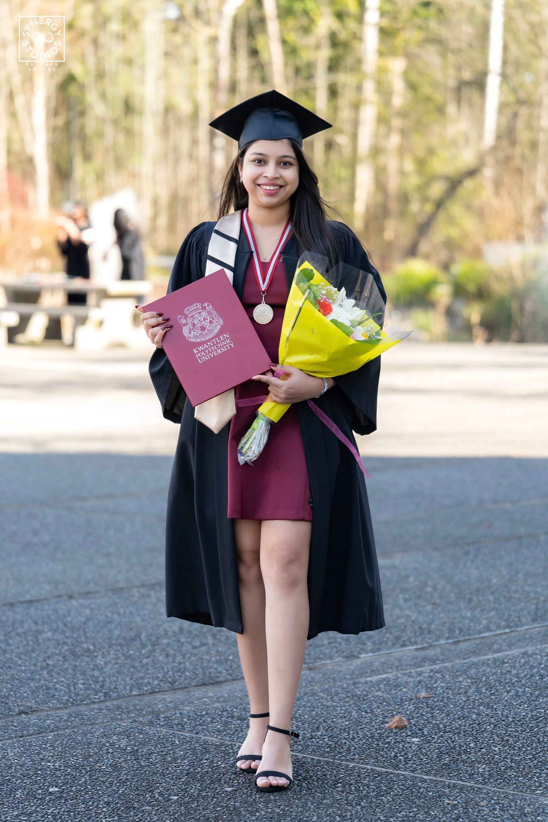 Graduate standing on a campus path with flowers and diploma