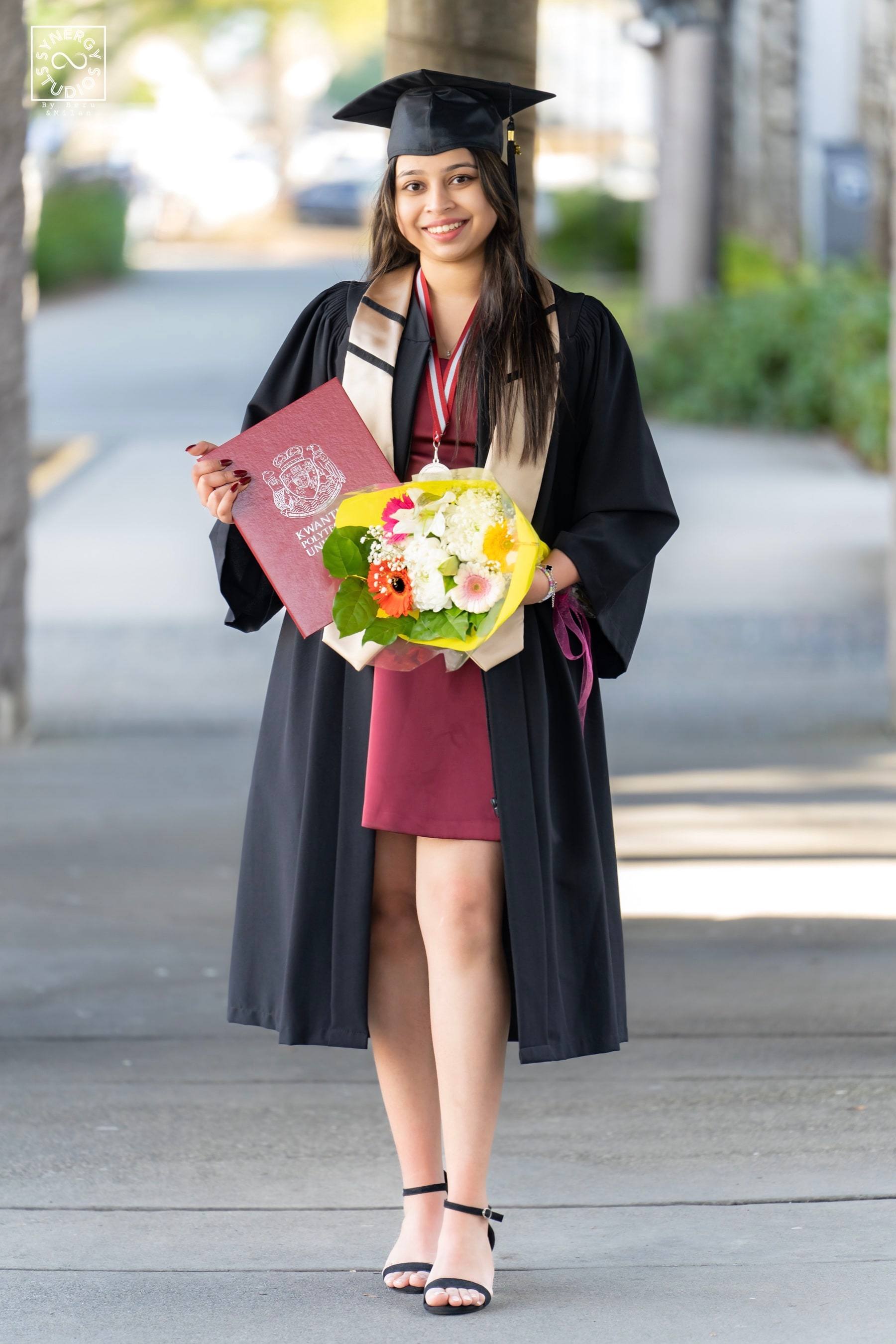 Graduate walking along a tree-lined campus path holding flowers